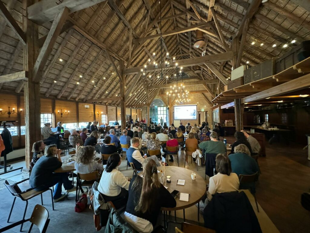 Ongeveer 80 mensen bijeen in De Kasteelboerderij van het Nederlands Openluchtmuseum. We zien mensen op de rug die aan tafels naar een presentatie voorin de zaal kijken, onder een gewelfd houten plafond met sfeerverlichting.